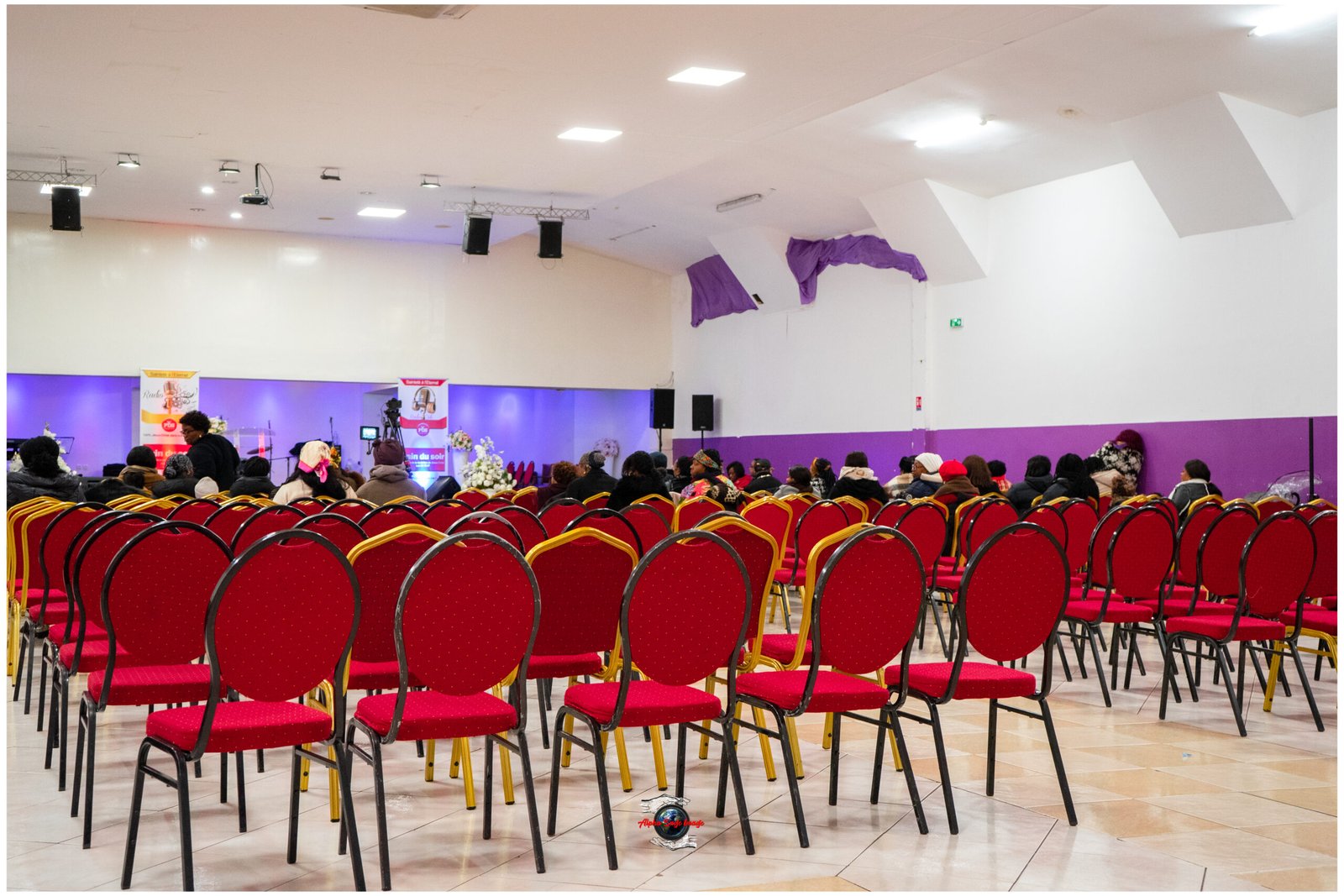 Audience seated in red chairs facing a small stage with banners in a brightly lit hall with purple accents.