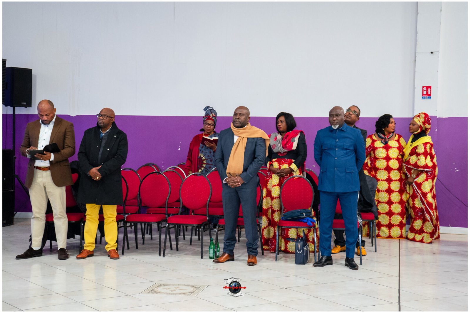 Group of adults in formal and traditional attire stand in a hall with red chairs and a purple-walled backdrop.