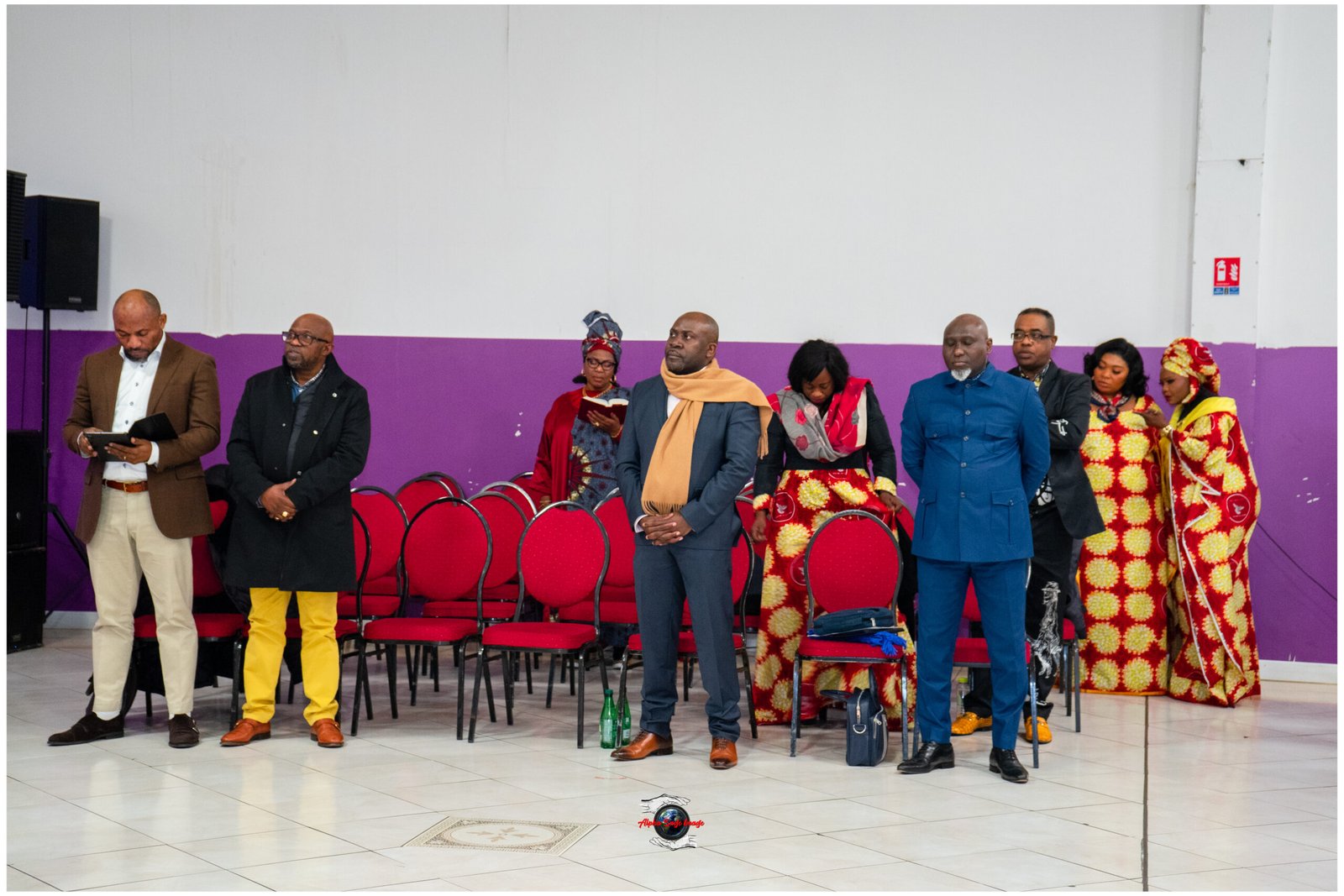 Group of casually dressed and traditionally clothed people standing in a row at an indoor event, with red chairs behind them and a purple wall backdrop.