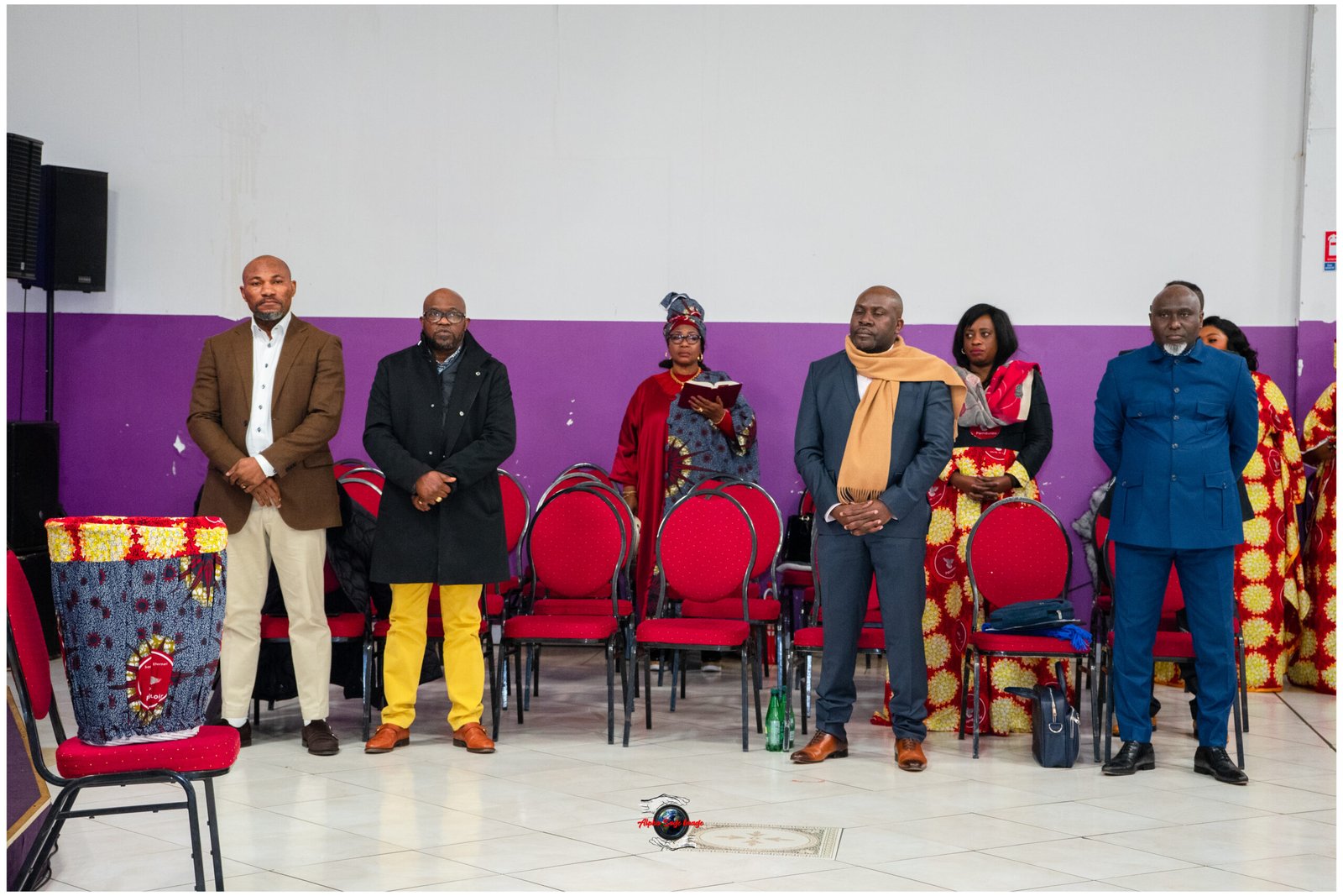 Seven adults stand in a row in a hall with a purple wall, some in suits and traditional attire, red chairs behind them.