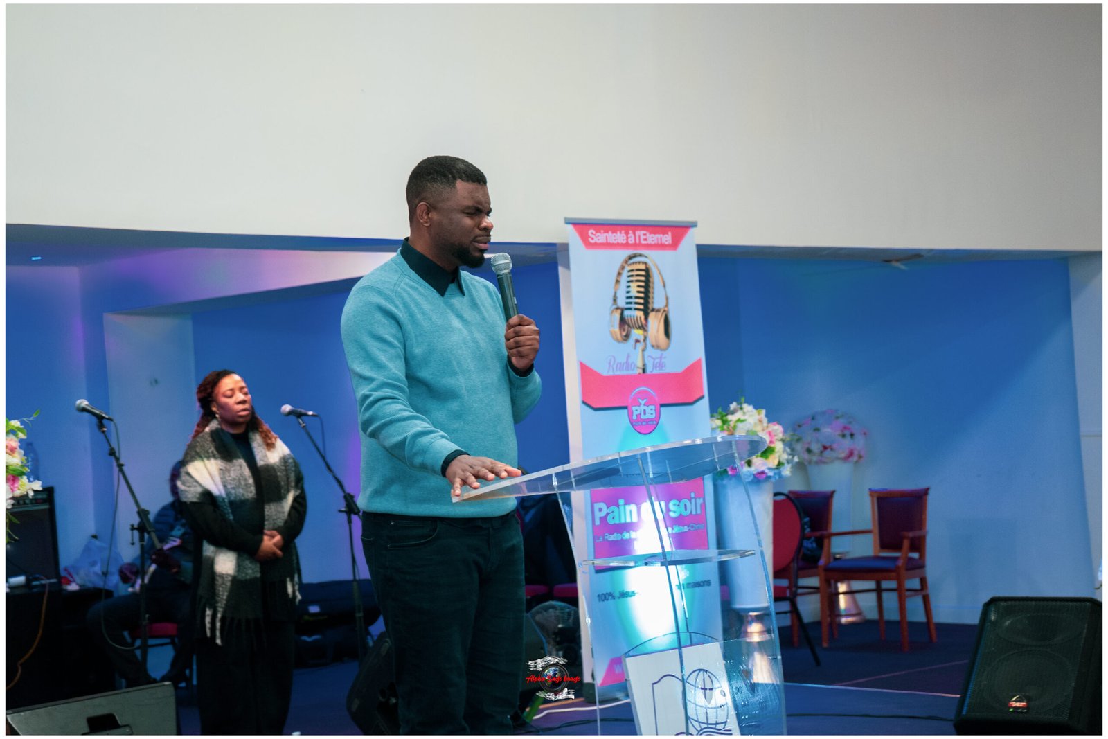 Man in a teal sweater speaks into a microphone at a clear podium on a blue-stage backdrop, with a woman and banner in the background.