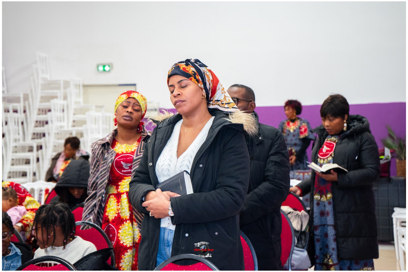 Group of people in colorful traditional clothing at an indoor event; woman in front with patterned headscarf holds a notebook.