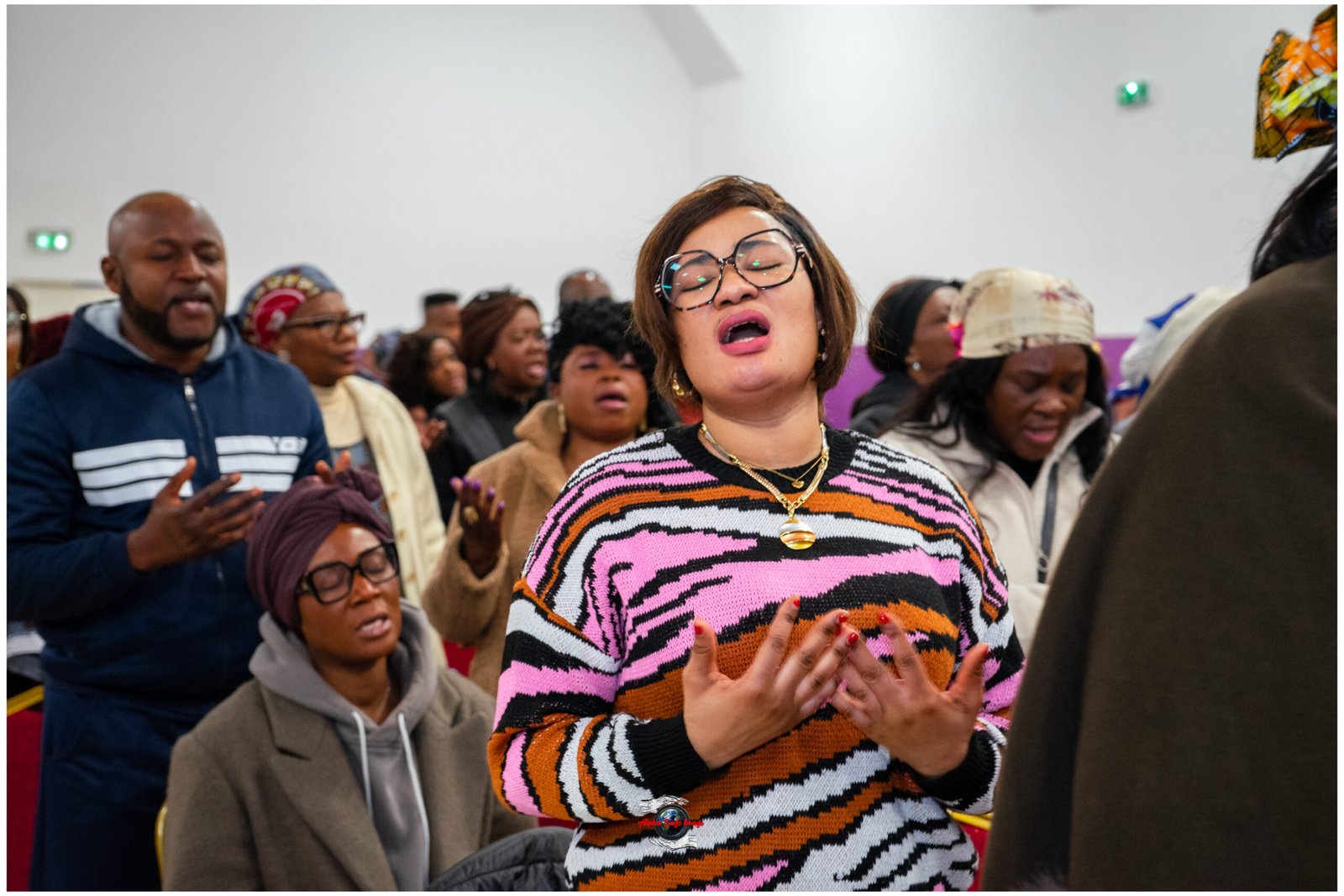 Group of people singing together at an indoor event; central woman with glasses and a bold pink‑orange striped sweater raises her hands in song.