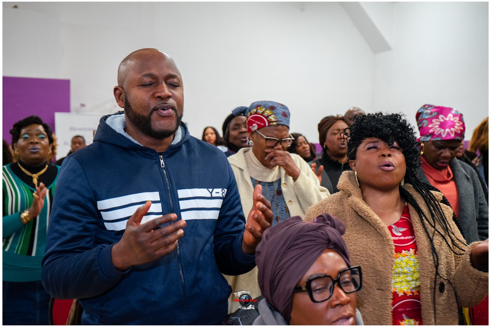 Group of people singing with hands raised in a bright room, wearing colorful outfits and head wraps.
