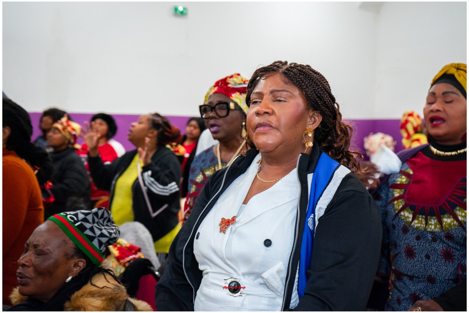 Group of women at a cultural gathering; foreground woman with braided hair in a white blouse and blue jacket, others in colorful head wraps behind her.