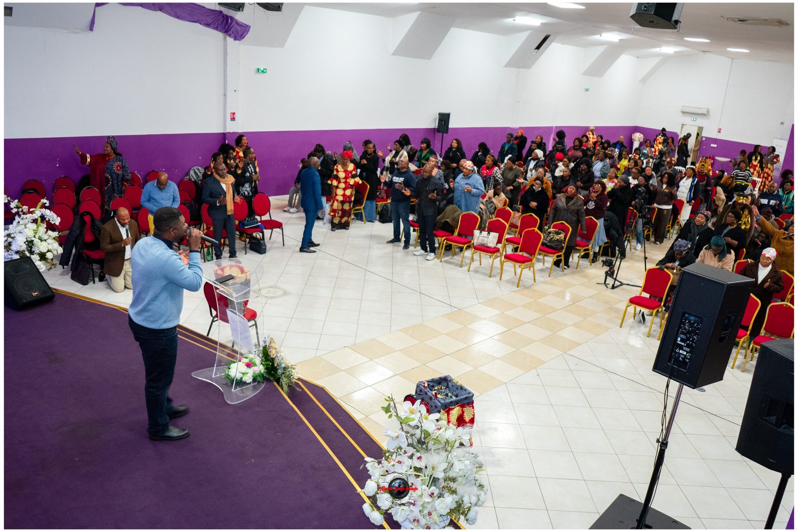 Man at clear podium speaking to a standing audience in a purple-walled hall with red chairs and floral decorations