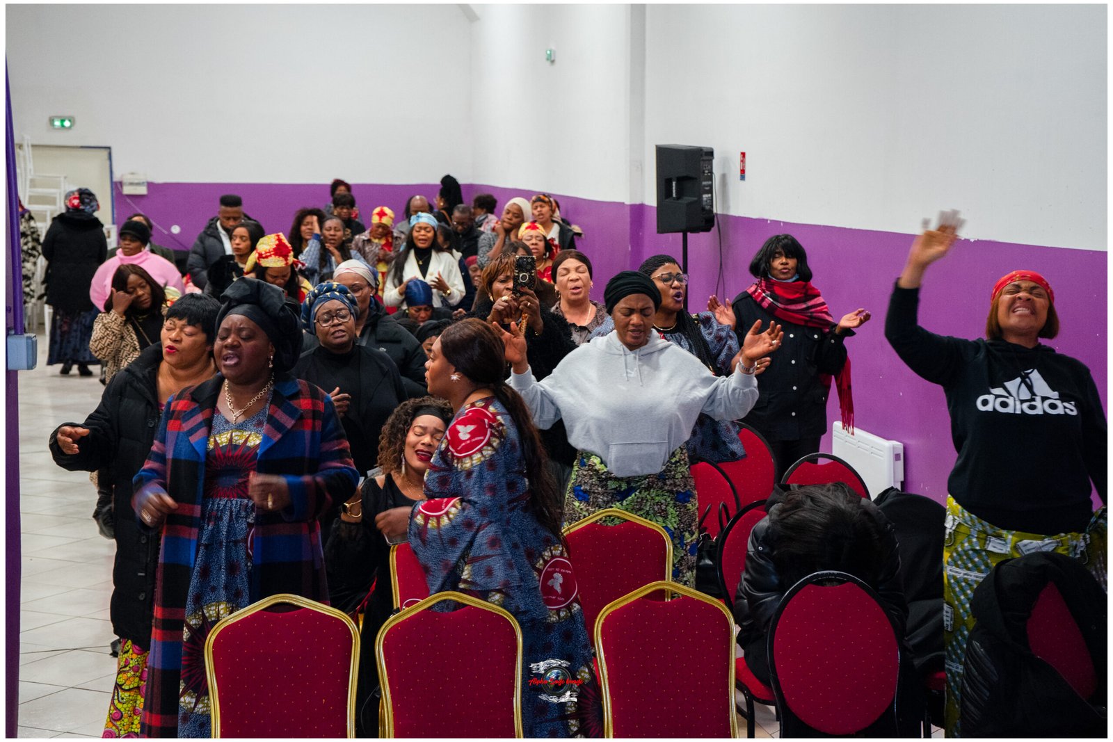 Crowd of women dancing and singing in a hall with purple wainscoting, many wearing colorful headwraps and patterned clothing.