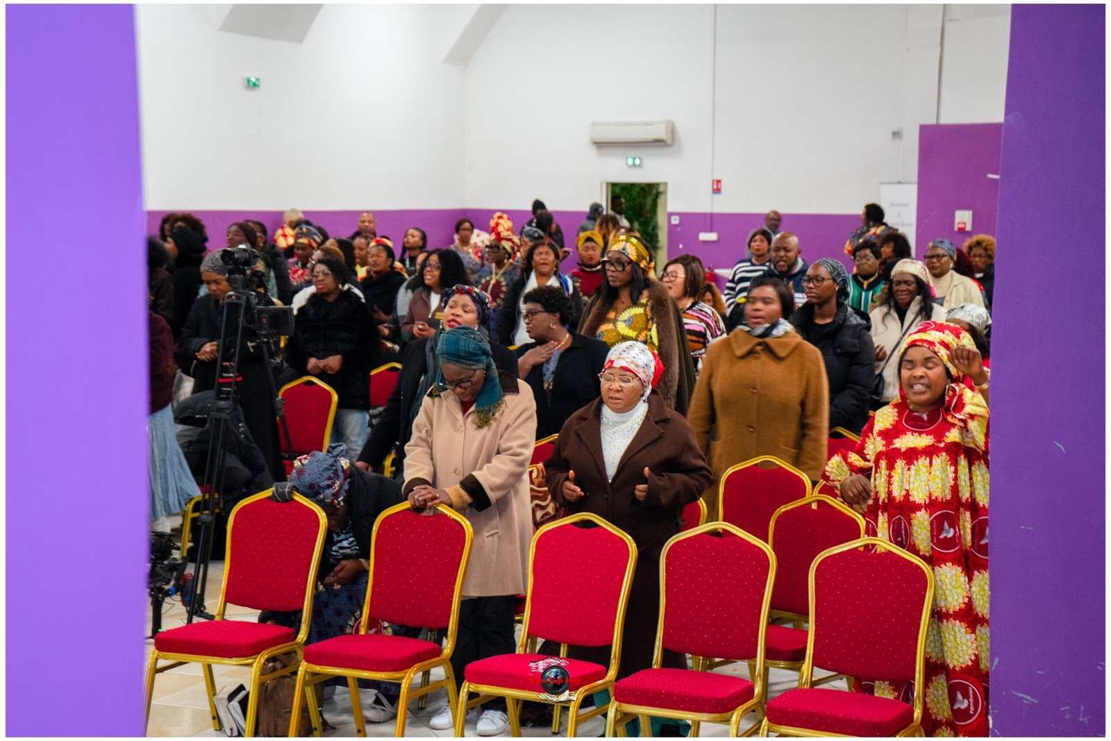Group of people standing and sitting at a cultural event in a hall with purple walls and red chairs.