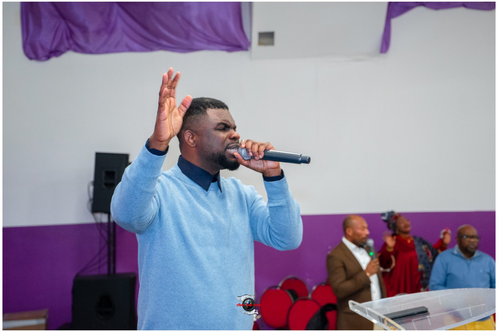 Man in a light blue sweater sings into a microphone on stage, with one hand raised, against a purple backdrop and other people in the background.