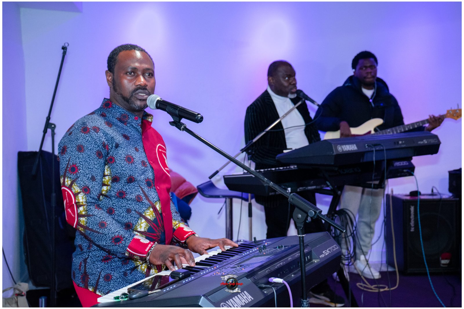 Frontman in a patterned shirt plays a Yamaha keyboard and sings into a microphone on a purple-lit stage, with two other musicians in the background.