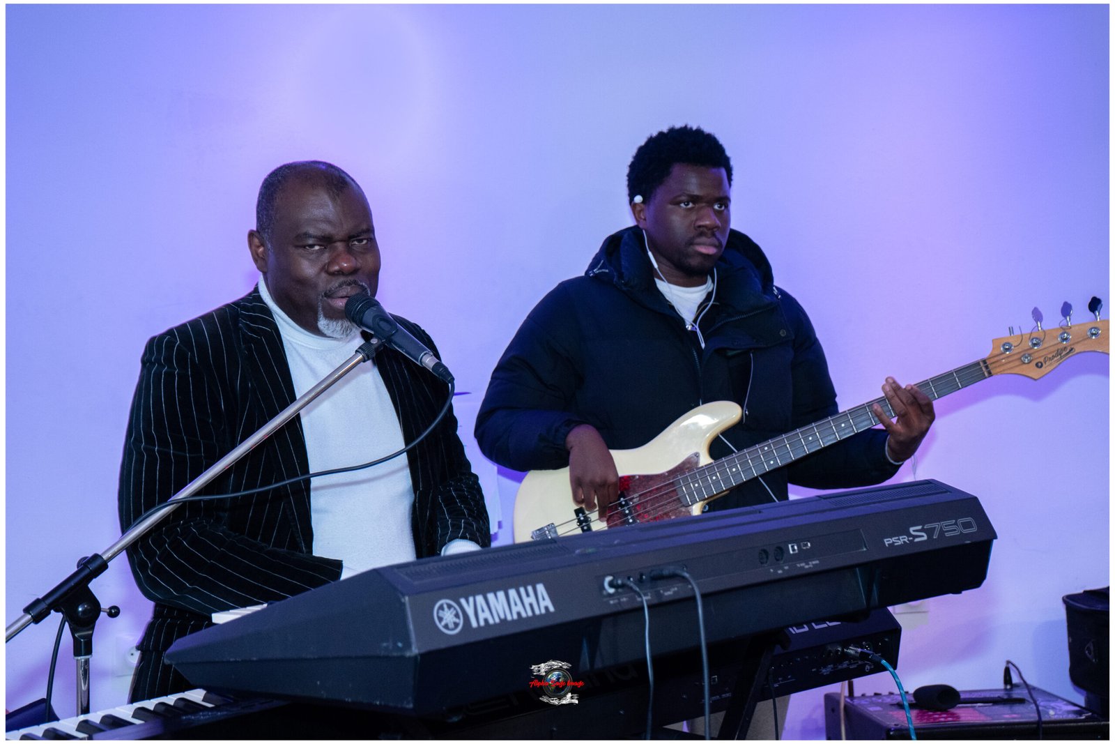 Two musicians performing on a blue-lit stage: a man at a Yamaha keyboard singing into a mic, and a bassist wearing a dark jacket beside him.