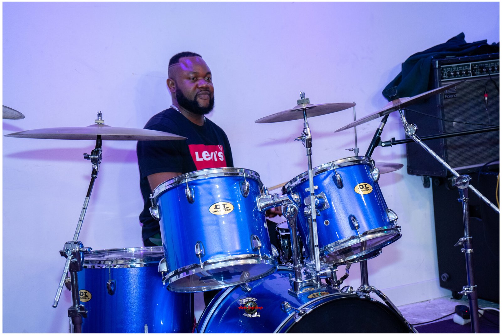 Drummer in a black Levi's shirt behind a bright blue drum kit on stage with cymbals nearby, looking to the left.