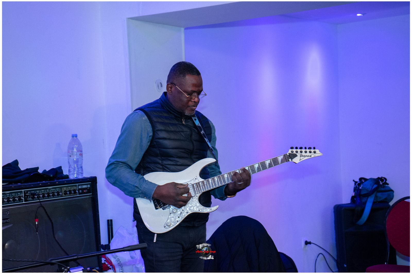 Man playing a white electric guitar in a blue-lit room beside a guitar amplifier and water bottle on a table.