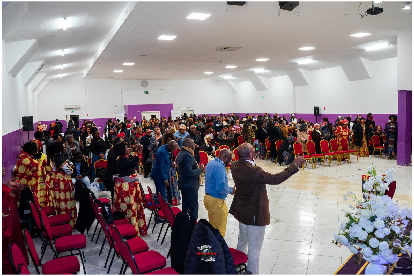 A crowded hall with people standing and seated, many wearing colorful traditional clothing, facing a stage area with purple walls and red chairs throughout.