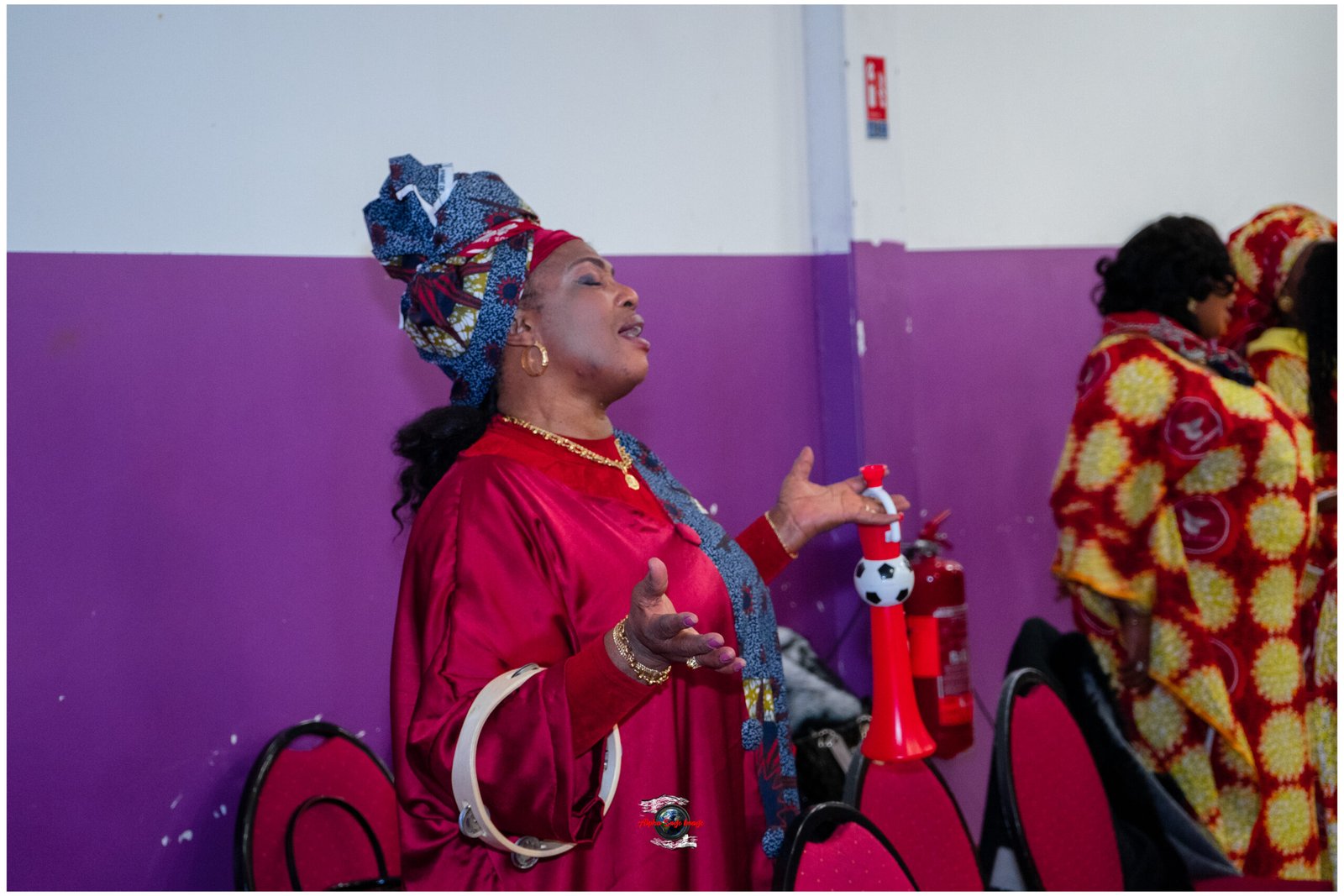 Woman in a bright red dress and patterned head wrap sings or speaks with hands raised, in front of a purple-walled room, others in colorful fabrics nearby.