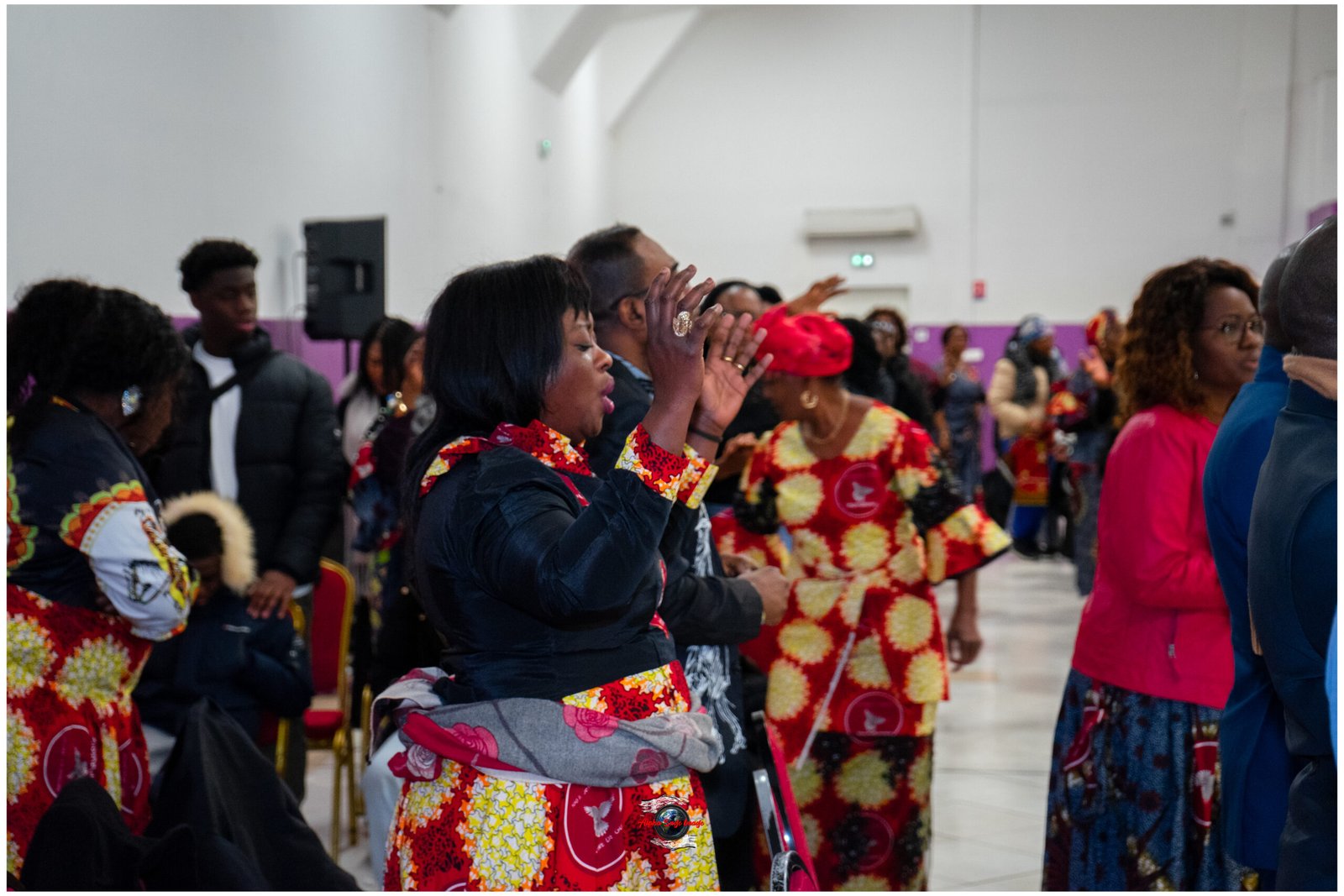 Group of people in a hall wearing colorful traditional African attire, clapping and singing together, foreground woman with hands raised in celebration.