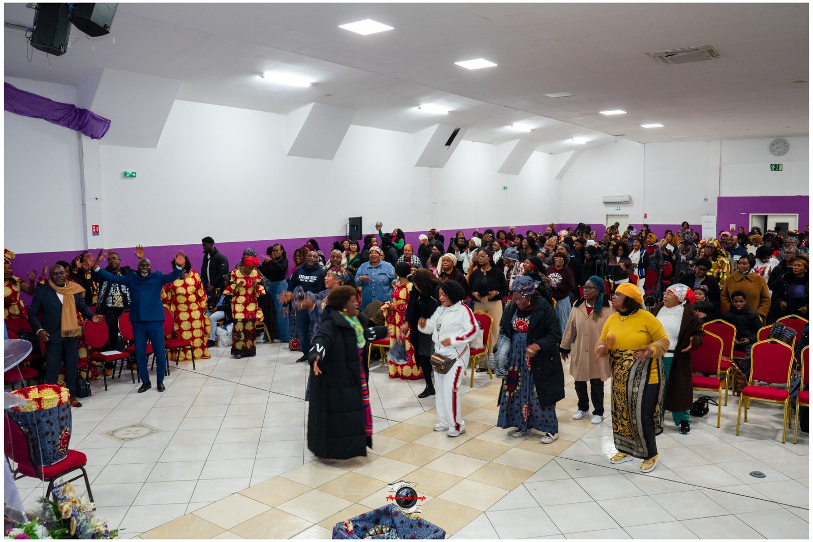 Large hall filled with people in colorful traditional clothing raising hands, mid-dance during a cultural event.