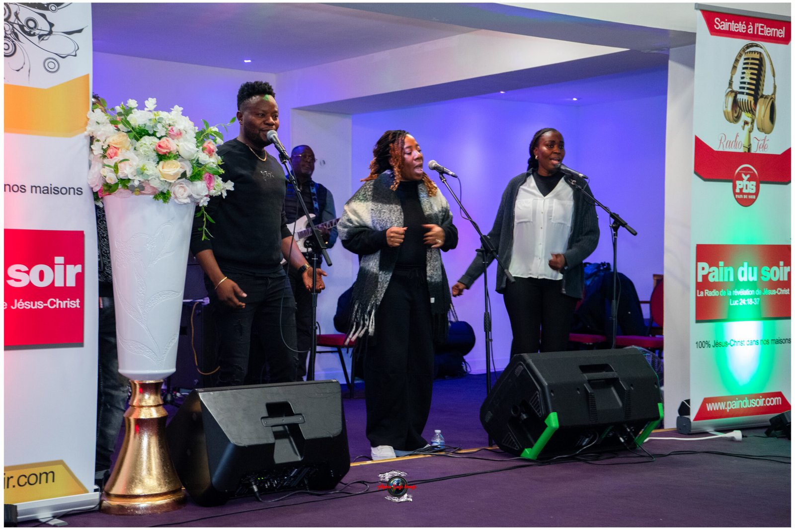 Three Black singers perform on a lit stage with a guitarist in the back and a tall flower arrangement on the left.