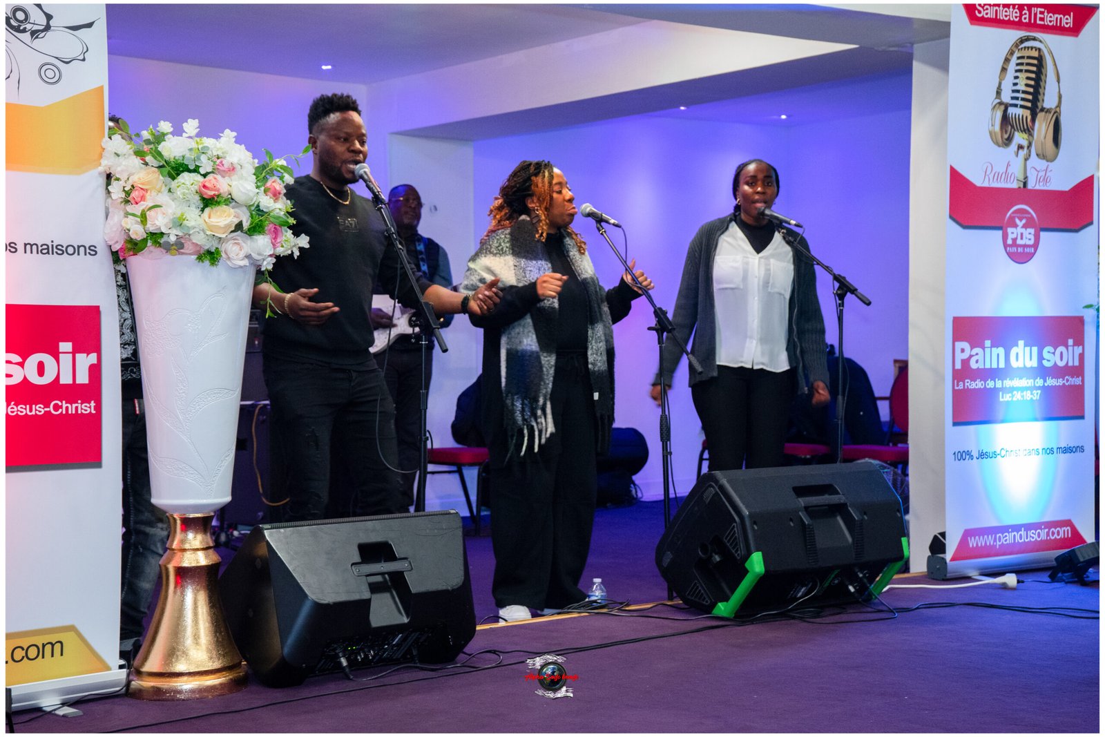 Three gospel singers perform on a lit stage with microphones, a guitarist in the back, and a tall white vase of roses on the left. Purple lighting and event banners are visible.