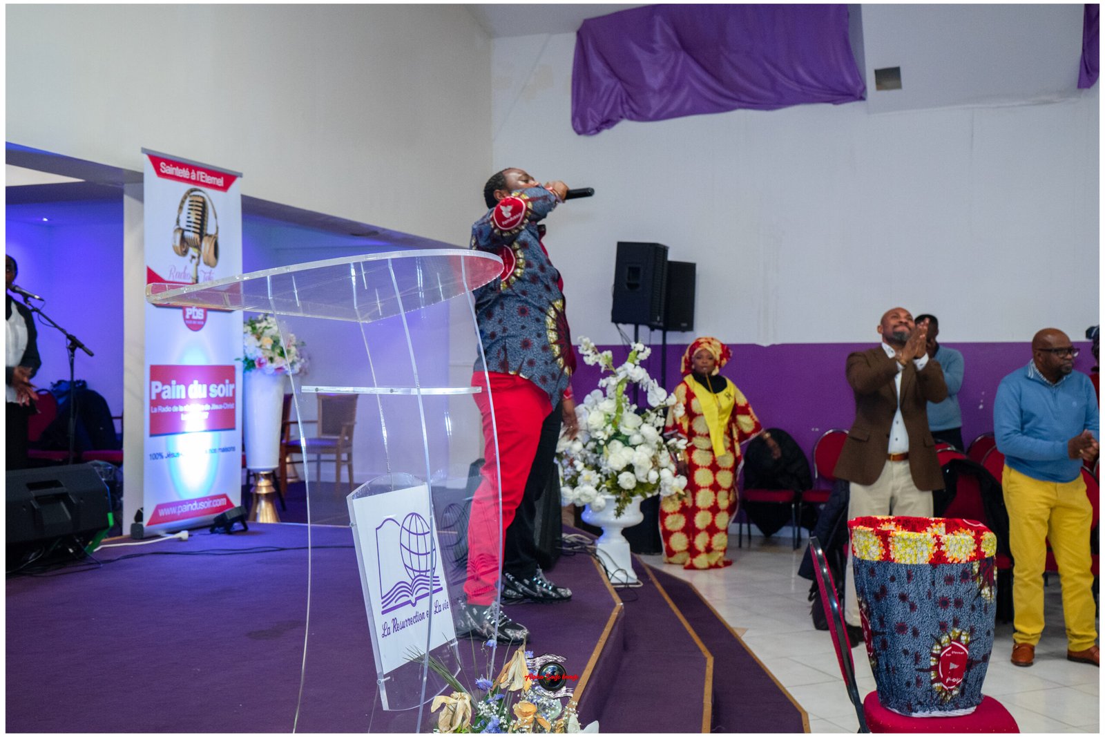 Man in colorful patterned shirt sings into microphone on a stage with a clear podium and purple decor, audience clapping in the background.