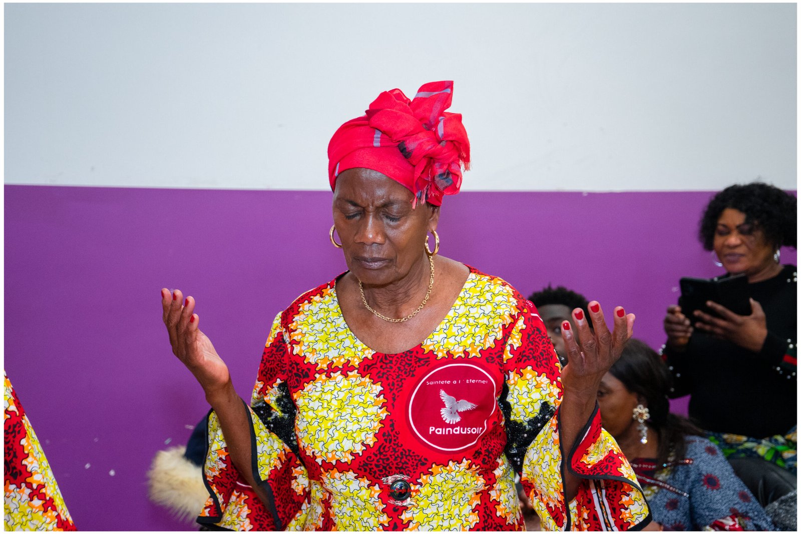 Woman wearing a red headwrap and colorful African-print dress with hands raised and eyes closed, likely dancing or praying against a purple backdrop.