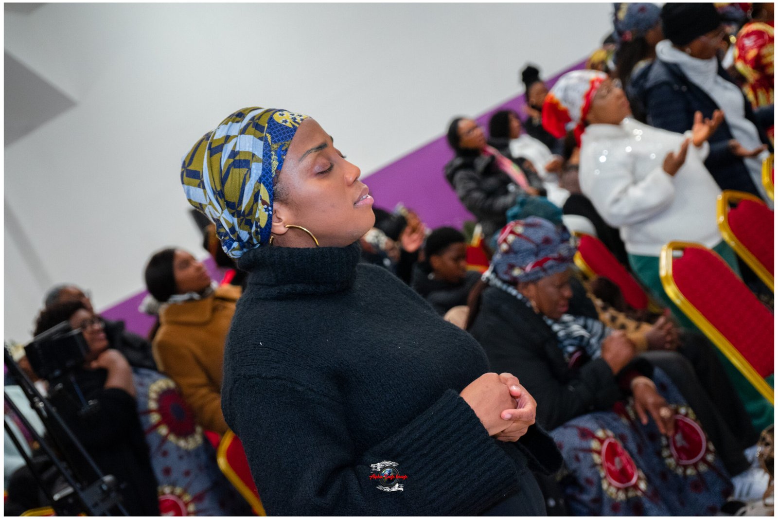 Foreground woman in a patterned blue-yellow head wrap, eyes closed, hands clasped in prayer at an indoor event with others seated in colorful chairs behind her.