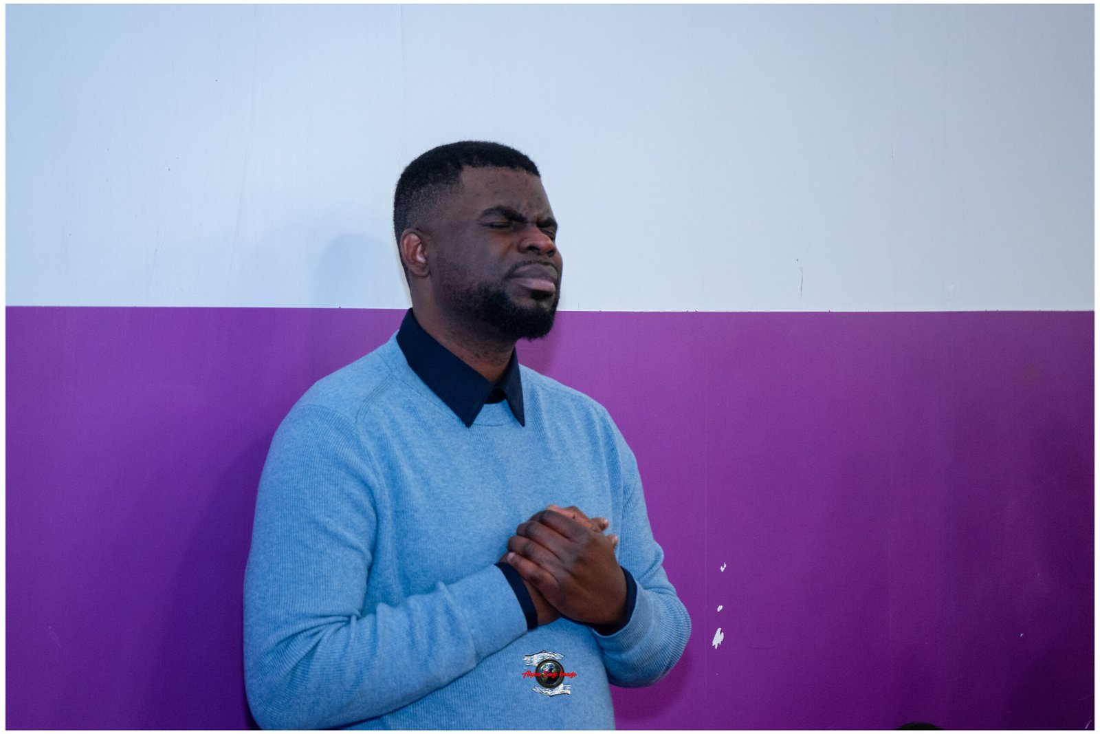Man with a short beard wearing a light blue sweater, eyes closed, hands folded in front against a two-tone blue-purple wall.