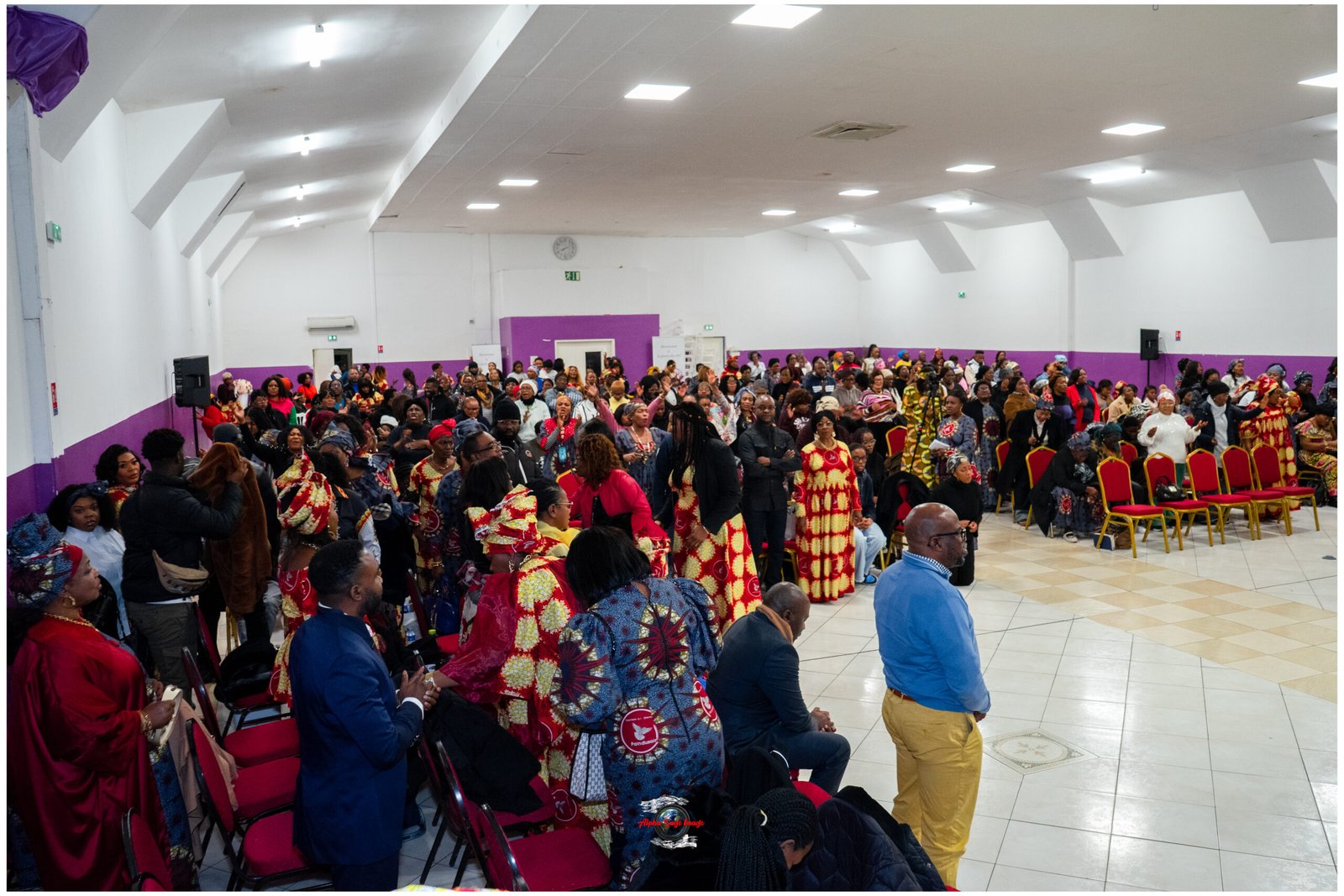 Large hall full of people wearing colorful traditional African outfits, many seated and others standing for a community event