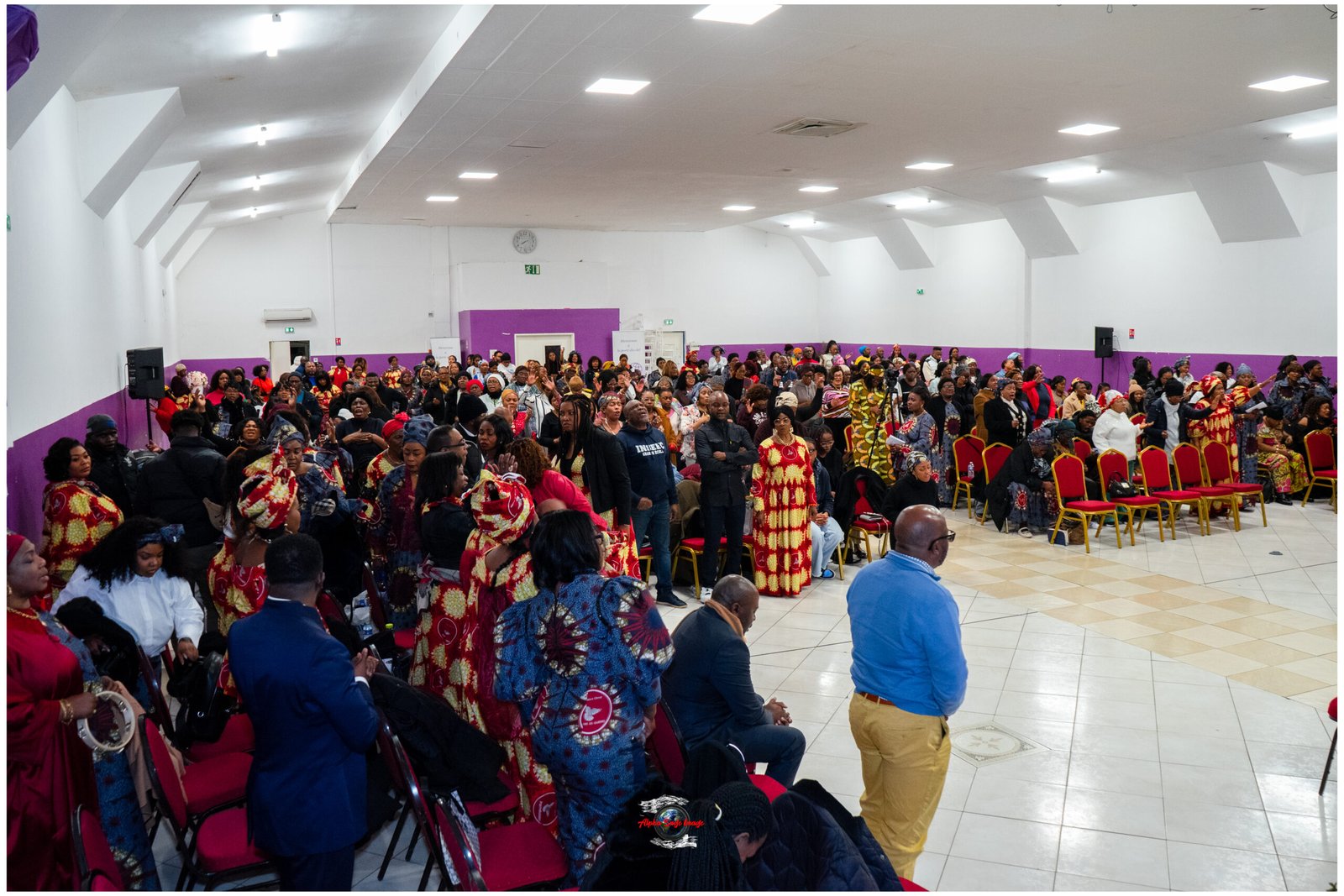Large hall filled with a crowded audience in vibrant traditional red and gold clothing, watching a cultural event.
