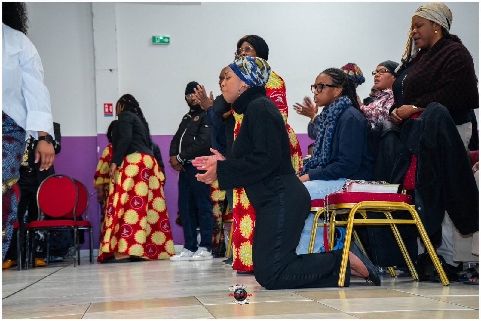 Group of people praying in a hall; woman in black kneels at center while others stand or sit, wearing colorful traditional dresses and head wraps.