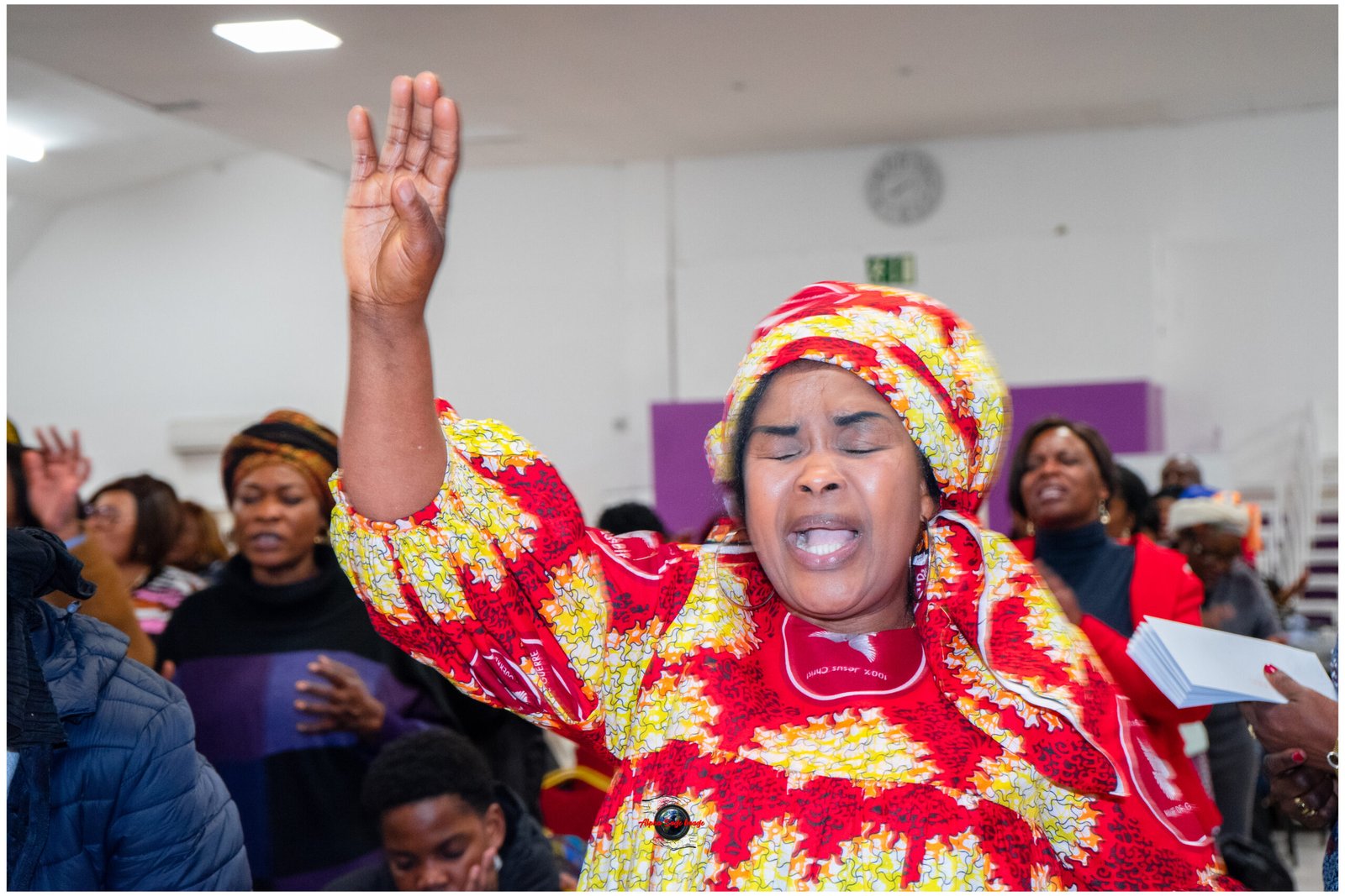 Woman in a bright red and yellow patterned dress raises her hand in celebration at an indoor gathering, with cheering people in the background.