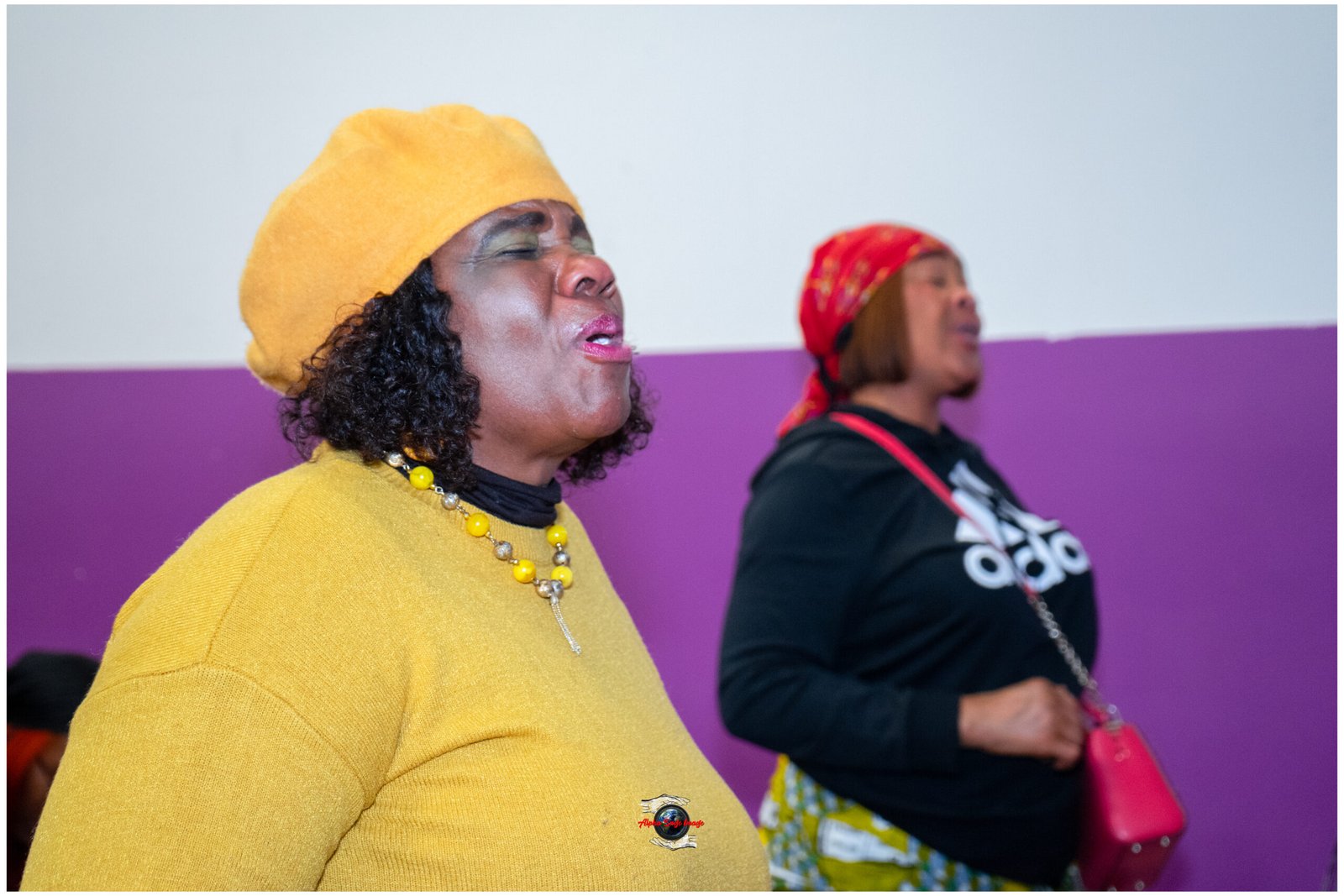 Close-up of a woman in a yellow hat and sweater singing with eyes closed, wearing a yellow bead necklace; a second woman in a red headscarf is blurred in the purple-background scene.