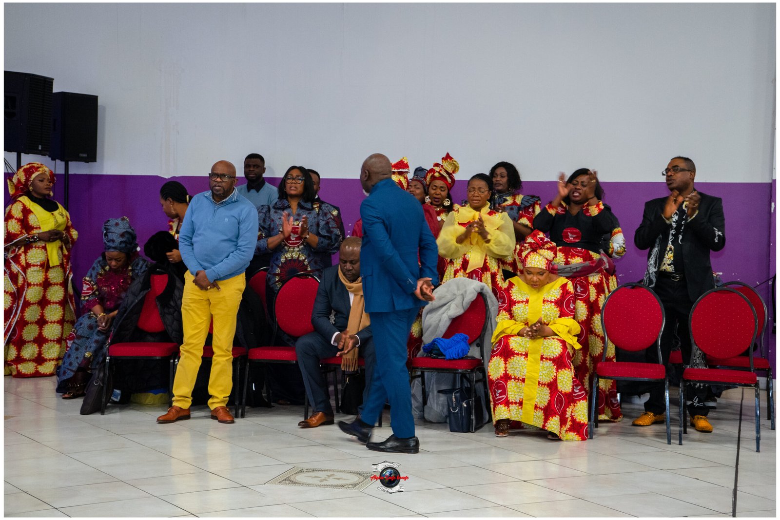 Group of people in bright traditional African outfits standing and clapping at a ceremony in a hall with a purple wall behind them.