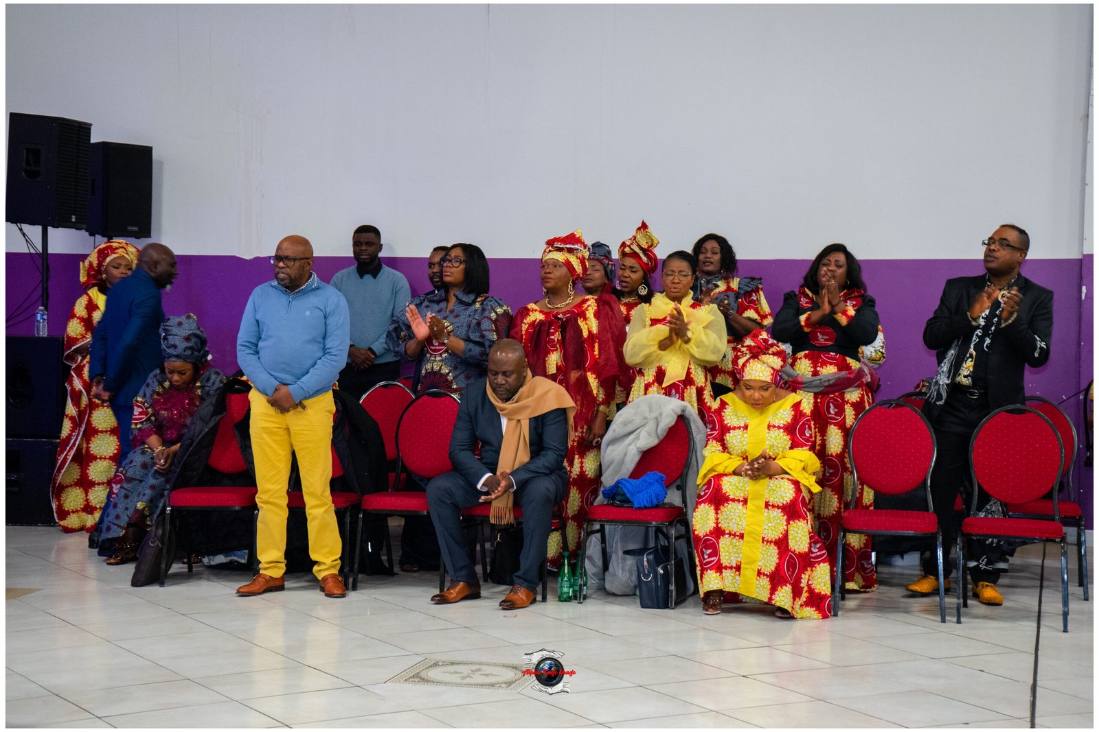 Group of people in bright red and yellow traditional attire standing and sitting in a hall with a purple wall behind them.