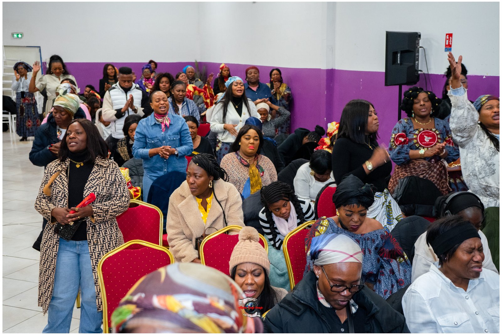 Group of women at a community or religious event, many seated on red chairs and some standing with hands raised, colorful traditional clothing, purple wall behind.