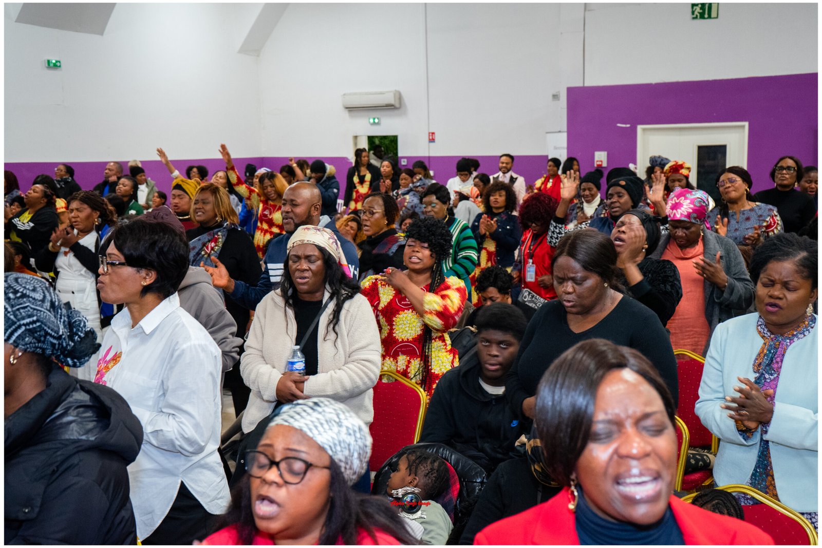 Large crowd of people in a hall with purple walls, many raising hands and singing or praying at a community event.