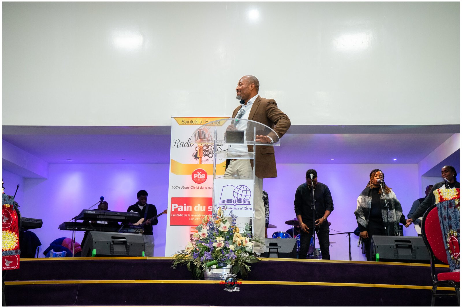 Man in a brown blazer speaking at a clear podium on a stage, with banners and a microphone, accompanied by musicians and floral arrangement in the foreground.
