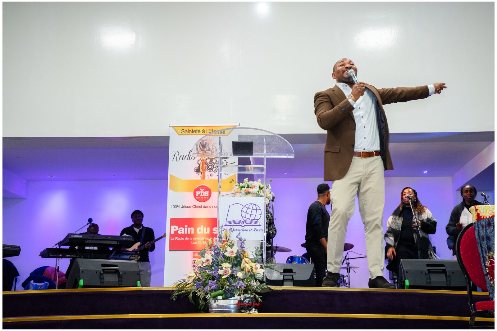 Male performer in a brown blazer sings into a handheld microphone on a stage, with a band and colorful banner behind him and floral arrangement in front.