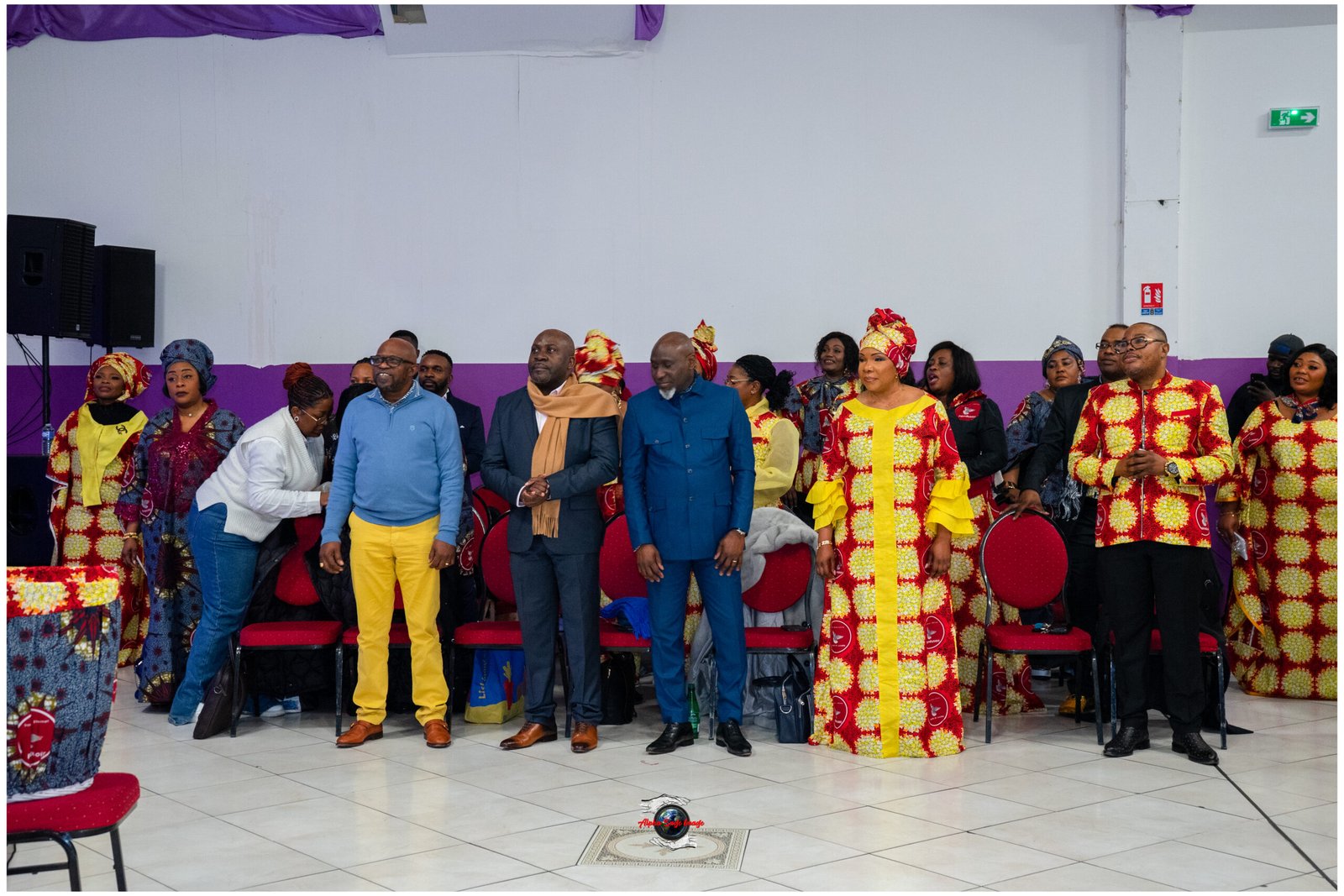 Group of people in vibrant African print attire standing in a hall with purple wall stripe and red chairs behind them.
