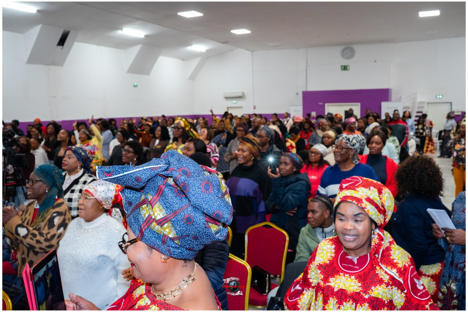 Large indoor gathering of predominantly African women in colorful traditional attire, seated and standing at an event.