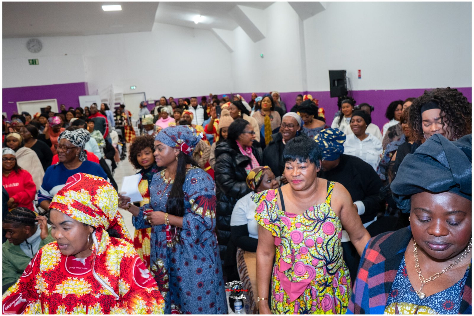 Crowd of people in bright traditional clothing gathered in a hall, many smiling and talking, with a purple wall in the background.