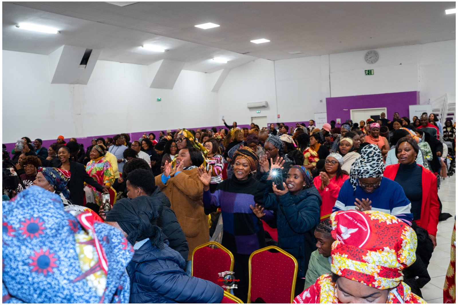 Large crowded event in a hall with purple walls; diverse audience wearing colorful clothing and headwraps, seated and standing.