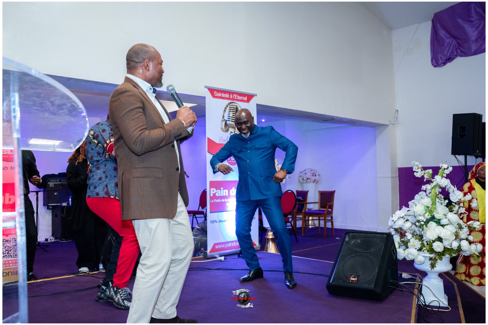 Man in a brown blazer speaks into a microphone on stage while a man in a blue suit dances nearby, during a church event with banners and flowers in the background