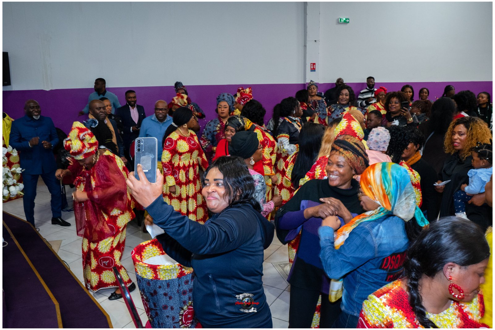 A lively crowd of people in bright patterned clothing dancing in a hall with a purple wall, some taking photos with smartphones.