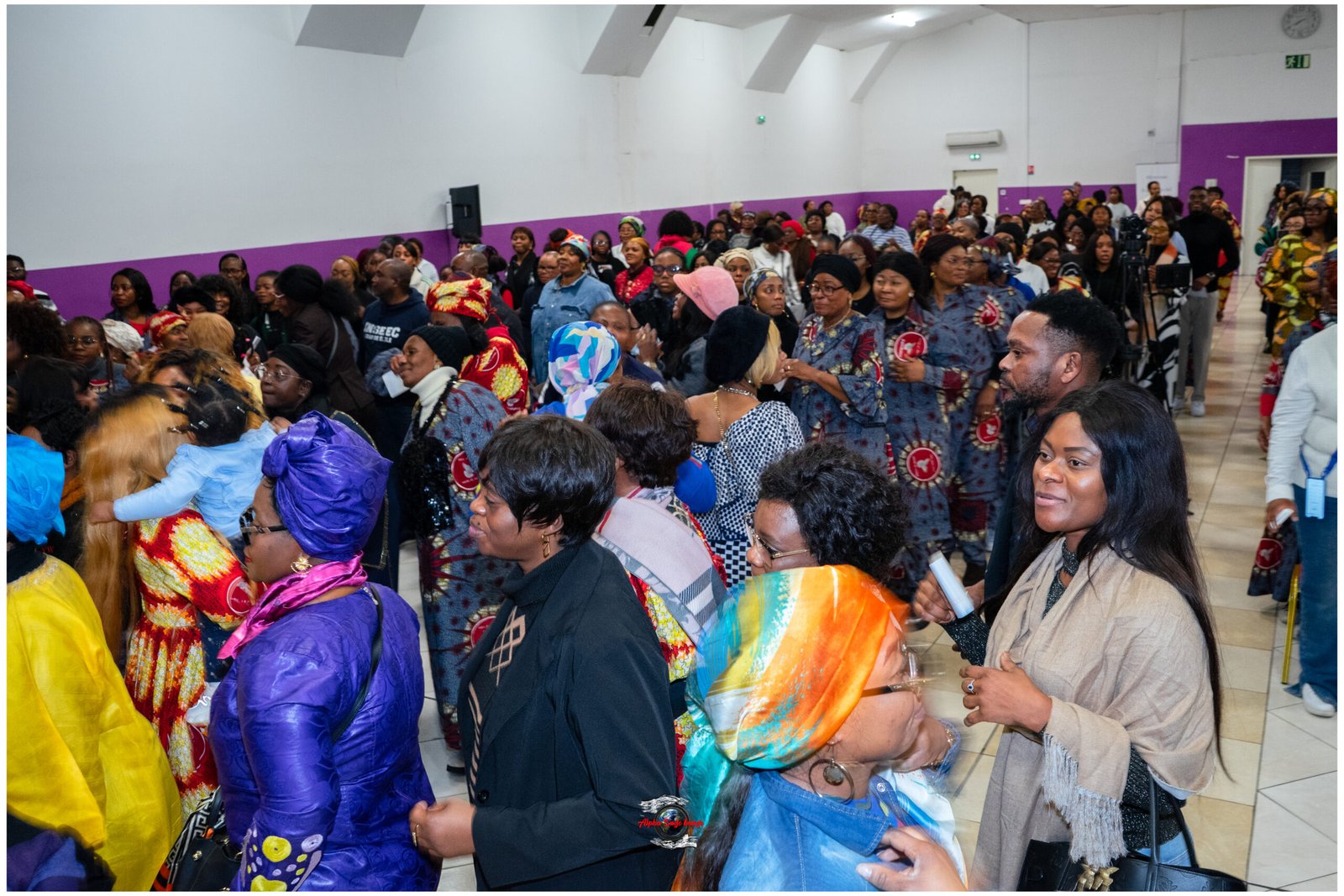 Crowd of people in colorful traditional outfits at an indoor community event. The hall has white walls with a purple stripe.