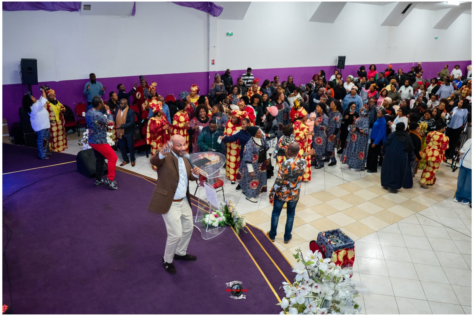Crowd of people in colorful traditional clothing at an indoor event, some dancing near a purple-carpeted stage with a speaker and microphone in the foreground.