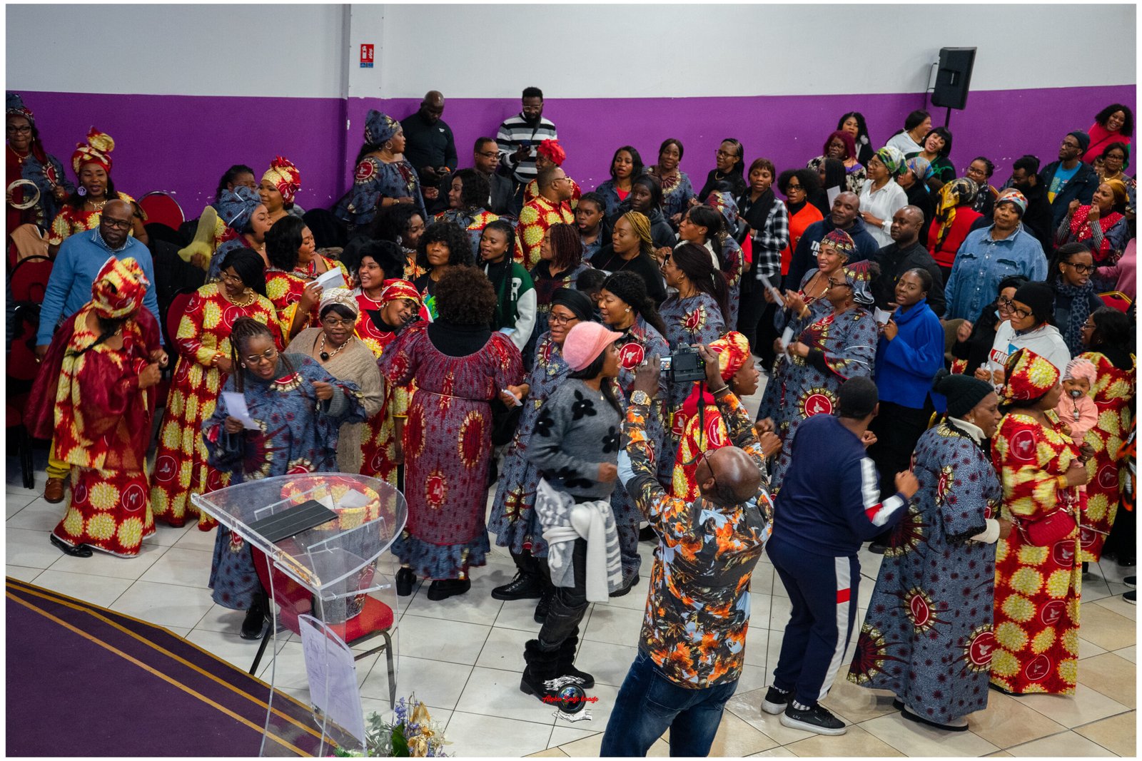 Large crowd of people in colorful African print dresses at an indoor cultural celebration.