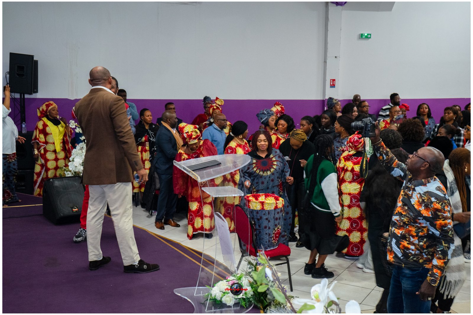 Audience in colorful traditional outfits gathers in an indoor venue around a clear podium and speaker in a brown blazer at the front.