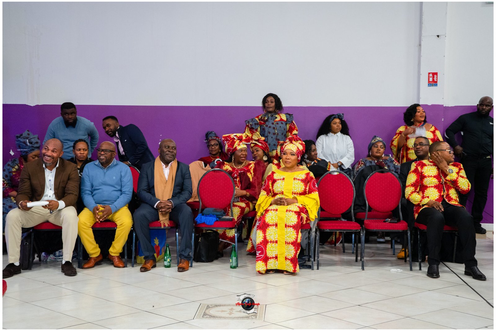 Row of people seated in a hall, with a woman in a bright yellow and red traditional dress standing center-stage against a purple wall backdrop.