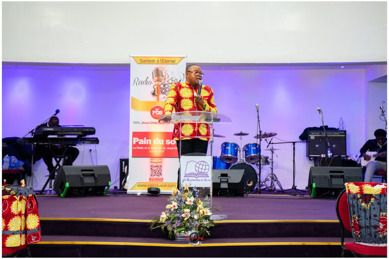 Man in a bright red and yellow patterned shirt speaks into a handheld microphone at a podium on a stage with a banner behind him and musical instruments nearby.